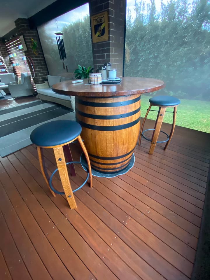A wooden barrel bar table with a round wood top and two matching wooden bar stools with dark blue seat cushions, set on a wooden deck.