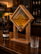 A wooden whiskey dispenser with a diamond-shaped glass reservoir and metal spout, shown on a dark wood bar counter beside a bottle of Glenfiddich 12 Year Old Scotch and a clear cut-glass tumbler. The glass reservoir is personalized with the name 'THE WILLIAMS BAR' and a crest.