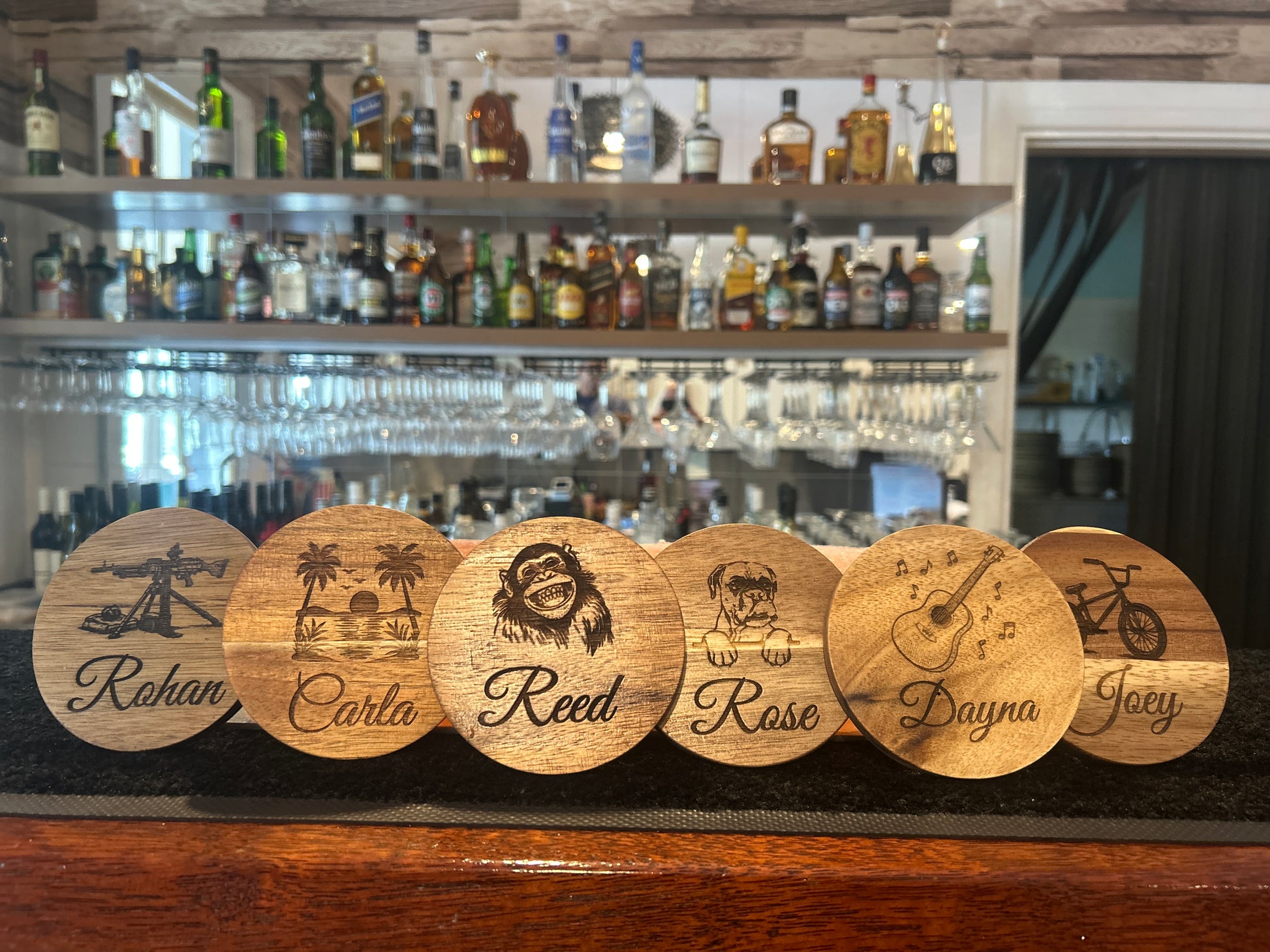 Wooden coasters with engraved names on a bar shelf with bottles and glasses in the background