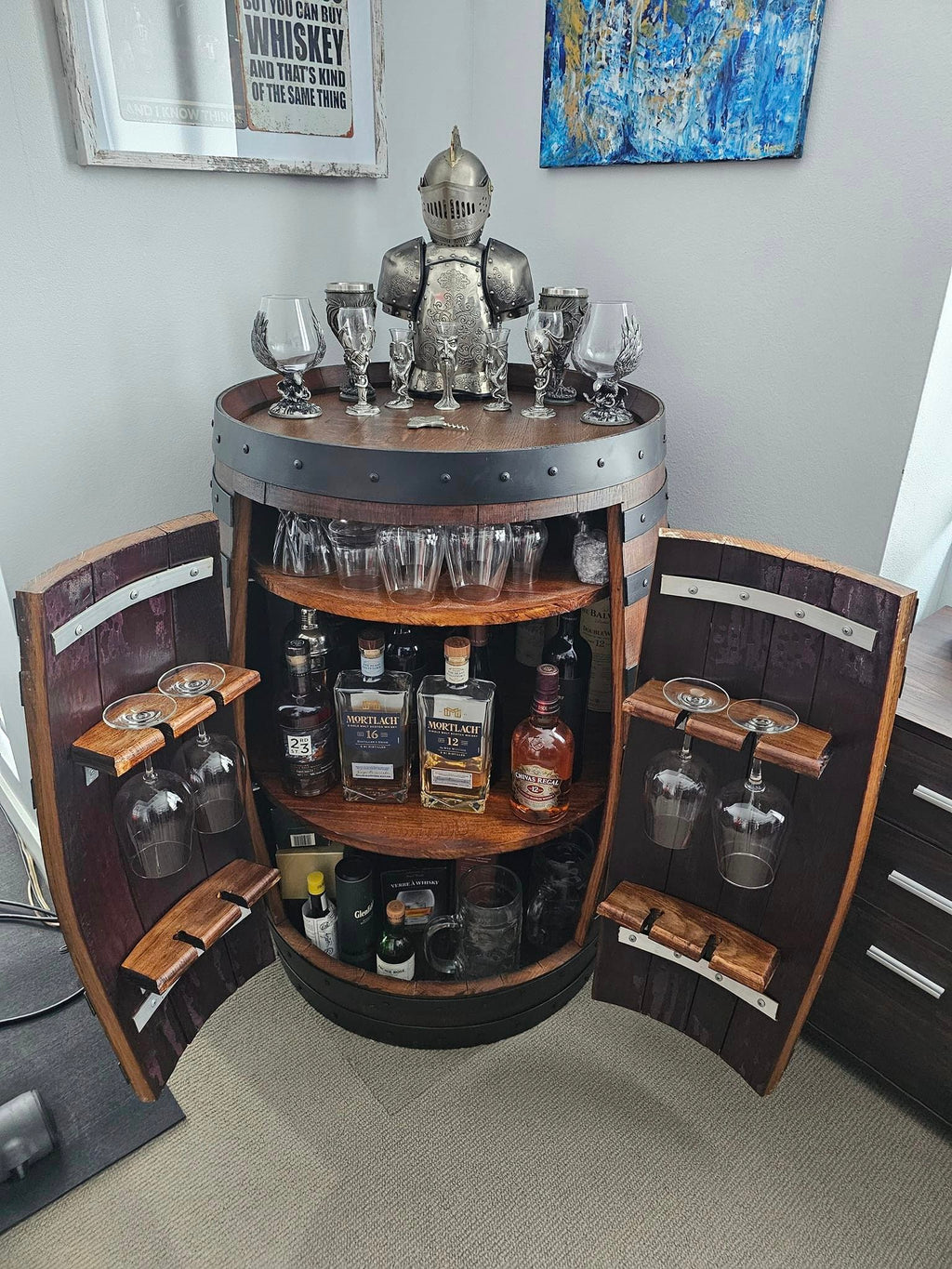 A rustic wine barrel barwith open side doors, made from dark wood and metal stonewash trim, holding liquor bottles, glasses, and a knight figurine on top, set in a room with a gray wall and framed art.